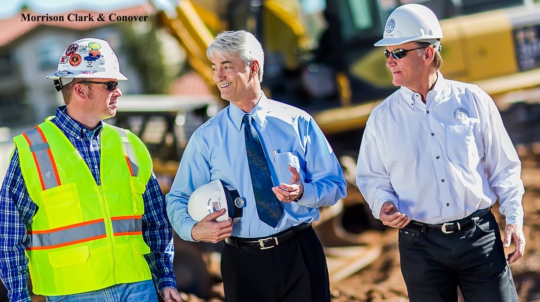 Three men in construction site, two wearing white hard hats and one wearing a high-visibility safety vest, engaged in conversation. The man in the middle is smiling and holding a white hard hat, while the other two men are listening.