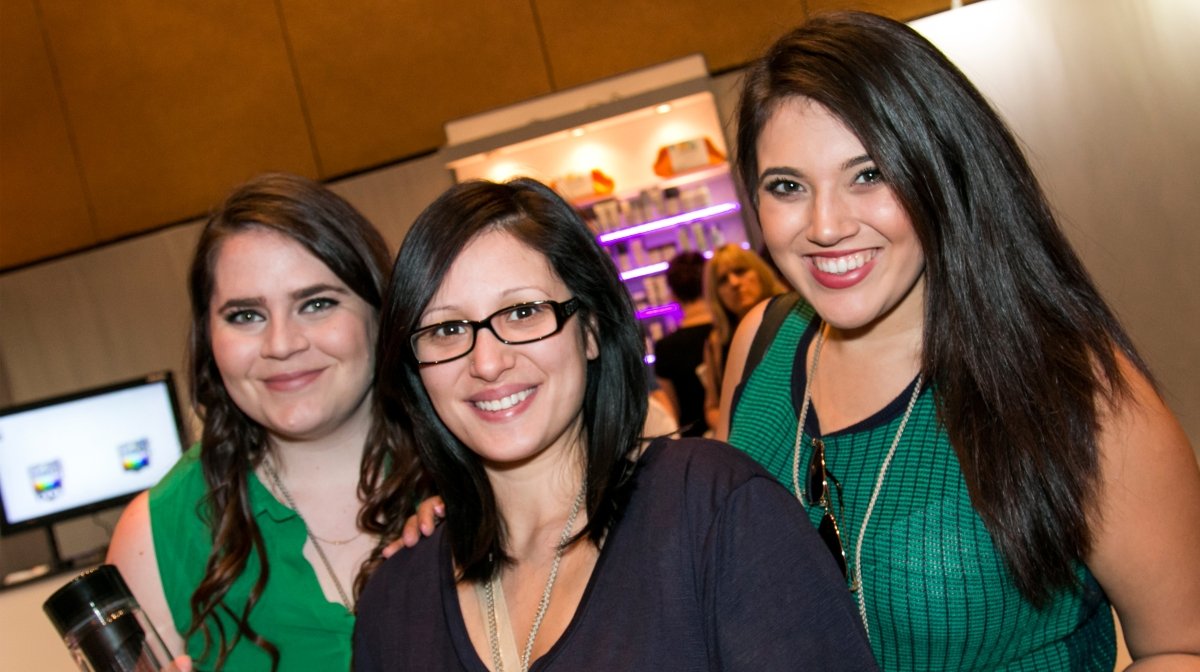 Three women smiling at a social event, standing close together indoors, with a bar and other guests visible in the background.