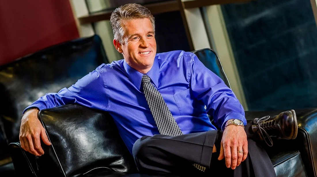 Smiling man in a blue shirt and patterned tie sitting on a black leather couch in a modern office setting.