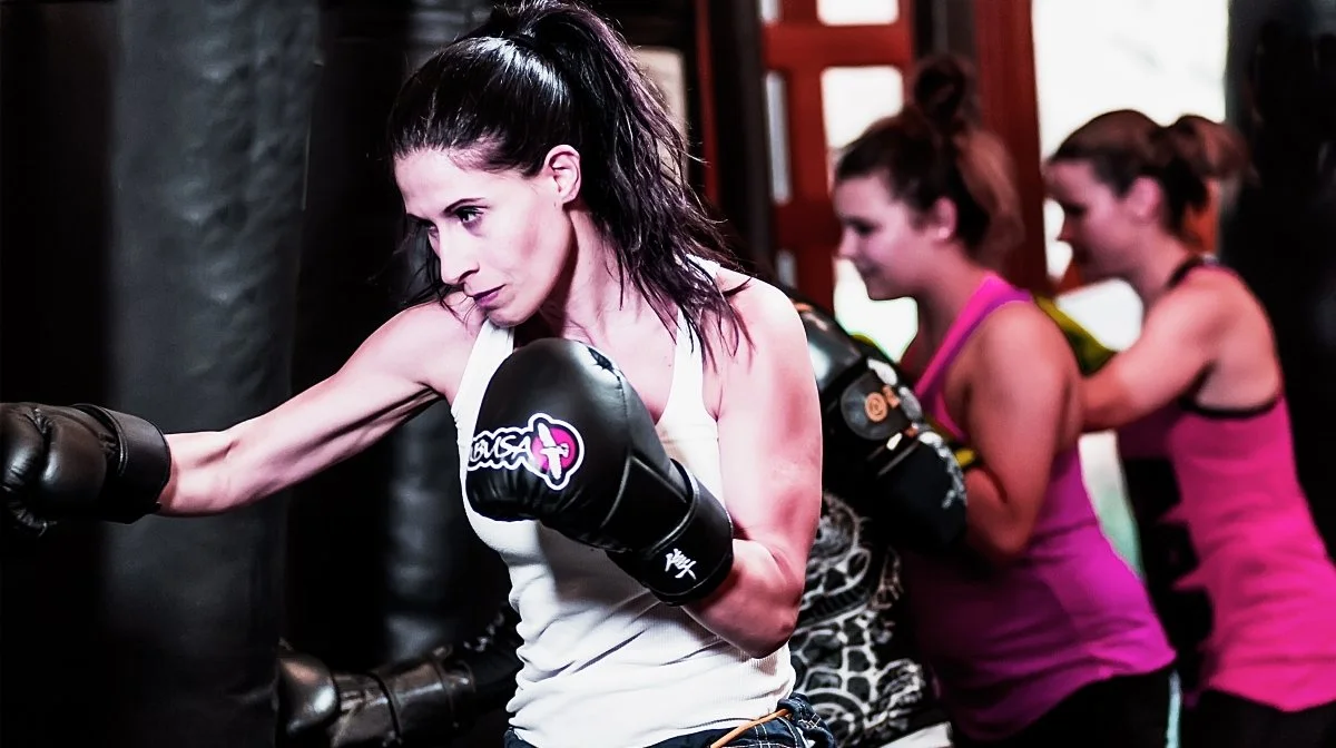 Women in a boxing gym practicing punches with a trainer, wearing athletic clothing and boxing gloves.