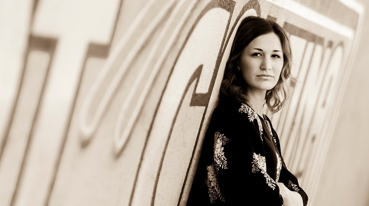 Young woman with wavy hair leaning against a wall with large letters, looking at the camera.