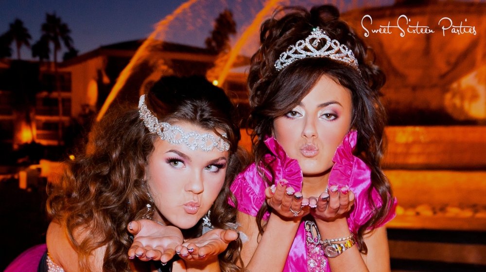 Two women wearing tiaras and pink dresses blowing kisses toward the camera at night, with a background of lit water fountains and buildings.