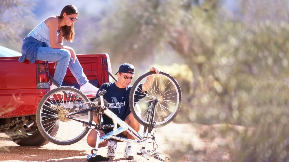 A woman sitting on the tailgate of a red pickup truck, watching a man in sunglasses and a cap fix a mountain bike on the ground in a desert-like area with trees in the background.