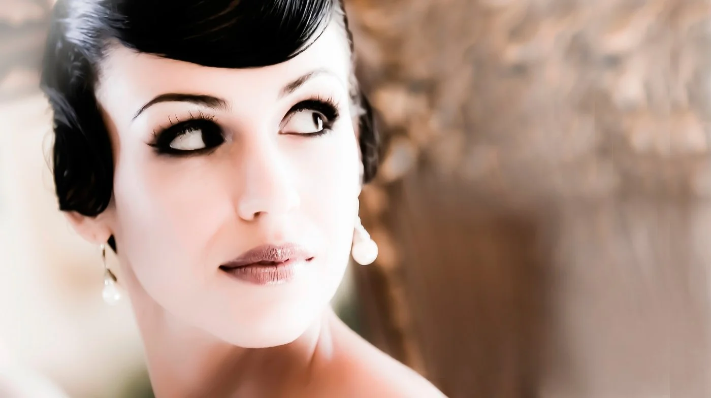 Close-up of a woman with dark, wavy hair and pearl earrings, looking to the side with a soft expression.