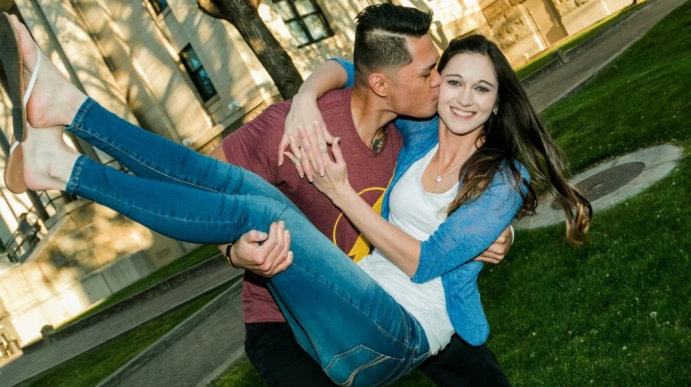 A man holding a woman in his arms while she smiles, outdoors in a park with trees and grass, and a building in the background.