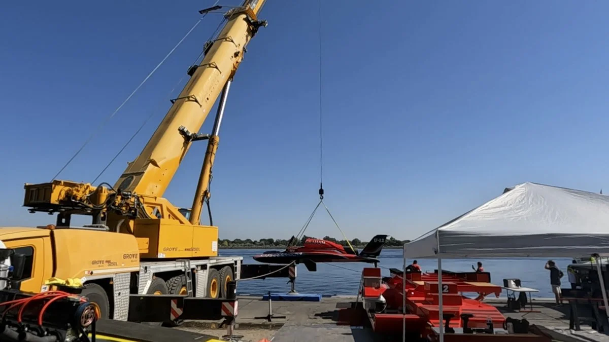 A yellow crane lifting a red and black boat out of the water near a white tent and red race boats by the river with a clear blue sky.