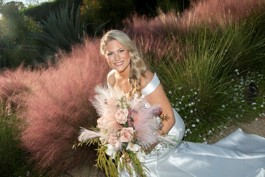 A smiling woman in a white wedding dress holding a bouquet of pink and white flowers seated among pink, green, and white ornamental grasses and plants.