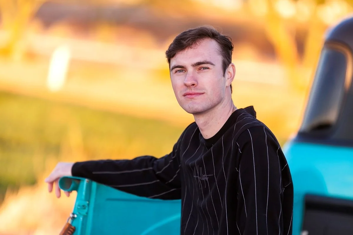 A young man with short brown hair and a slight smile, wearing a black shirt with white pinstripes, sitting outdoors next to a teal vehicle during sunset with a blurred background of trees and grass.