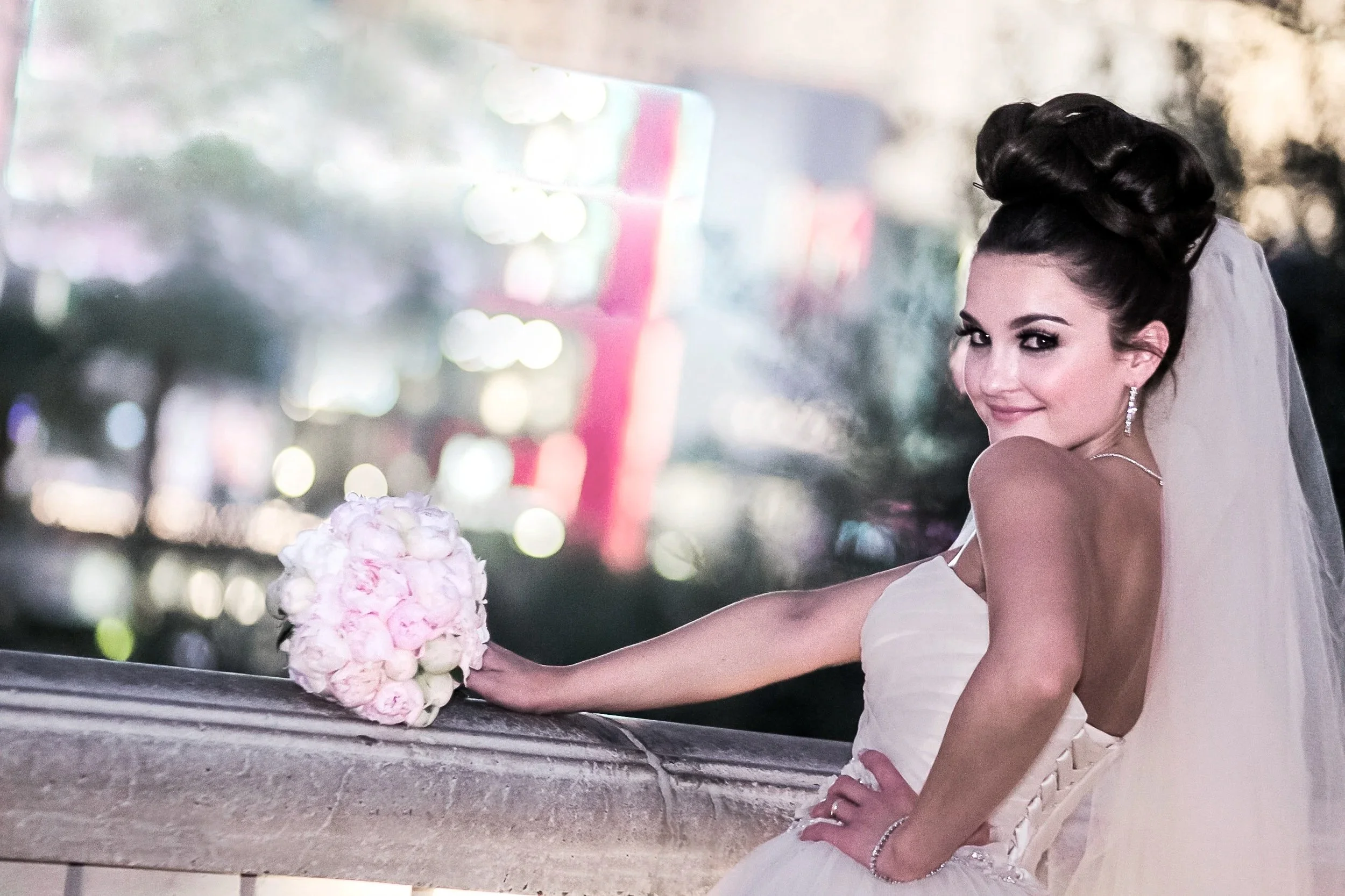 A bride in a white wedding dress with a veil, smiling and posing with her hand on her hip, holding a bouquet of pink and white flowers, on an outdoor balcony at dusk with blurred city lights in the background.