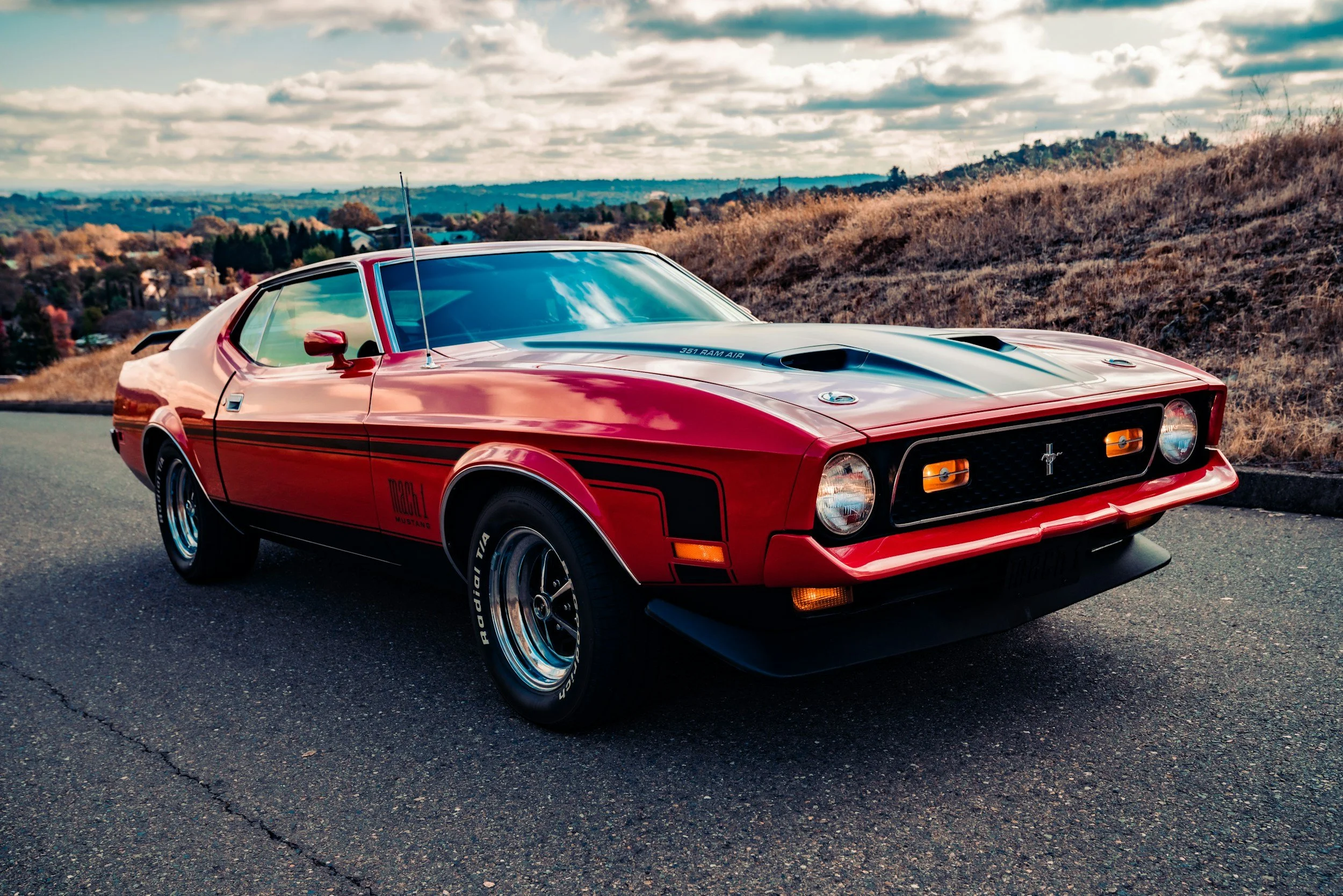 A red vintage Ford Mustang car parked on the side of a road with a scenic landscape of rolling hills and a cloudy sky in the background.