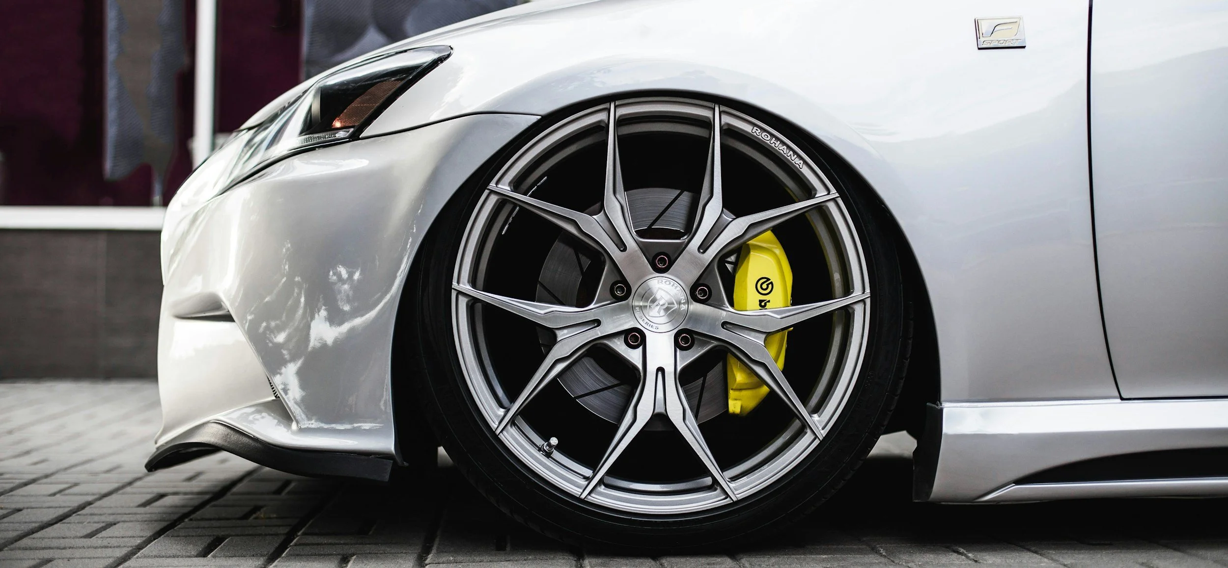 Close-up of a silver luxury car's front wheel with a custom alloy rim, a yellow brake caliper, and a black brake disc, parked on a paved surface.