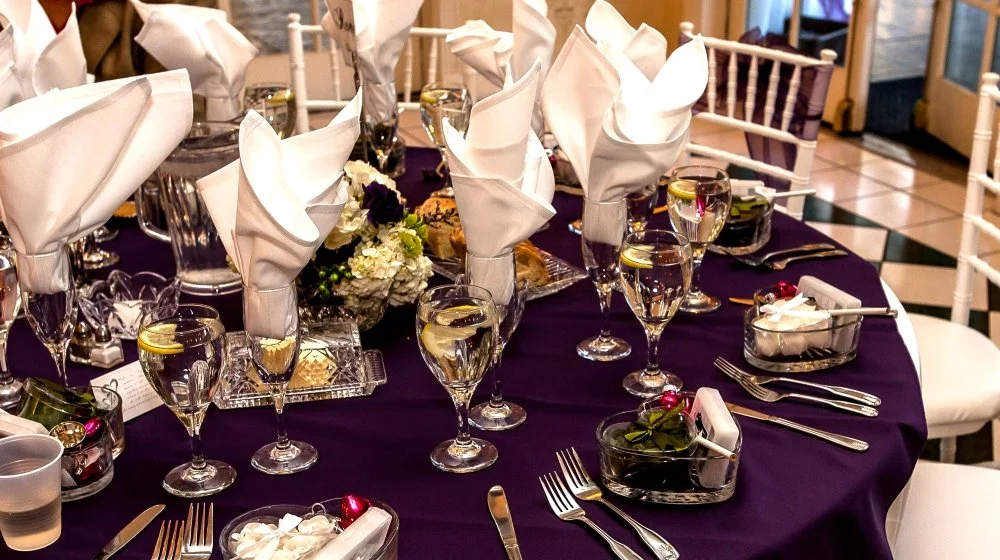 A table set for a formal dinner with white napkins folded like flowers, wine glasses filled with white wine or water, silverware, and small decorative bowls with cherries and other items in a restaurant.