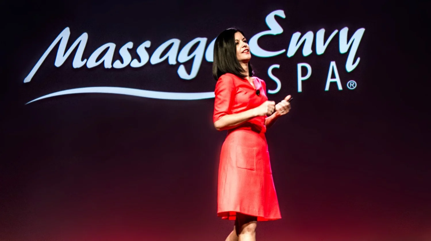 A woman in a red dress is speaking or presenting on stage at a spa conference or event. Behind her, there is a large sign that reads 'Massage Envy SPA' with a stylized script and logo.