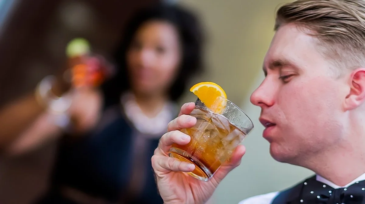 A young man in a tuxedo drinks a glass of cocktail with a lemon wedge garnish, at a social gathering with two blurred women in the background holding drinks.