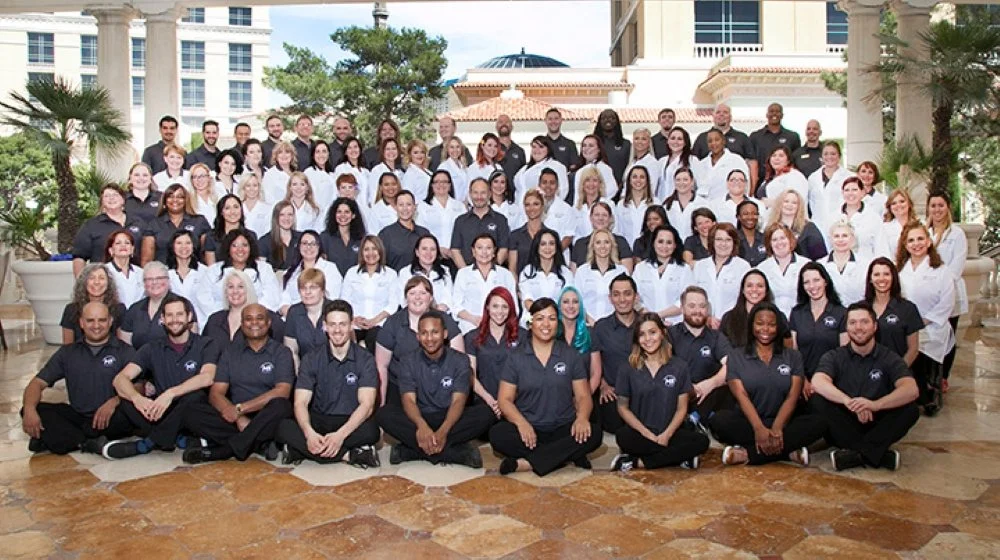 Large group photo of men and women in business attire, indoors, with some wearing white shirts and others in dark shirts, in front of a building with trees and a balcony.