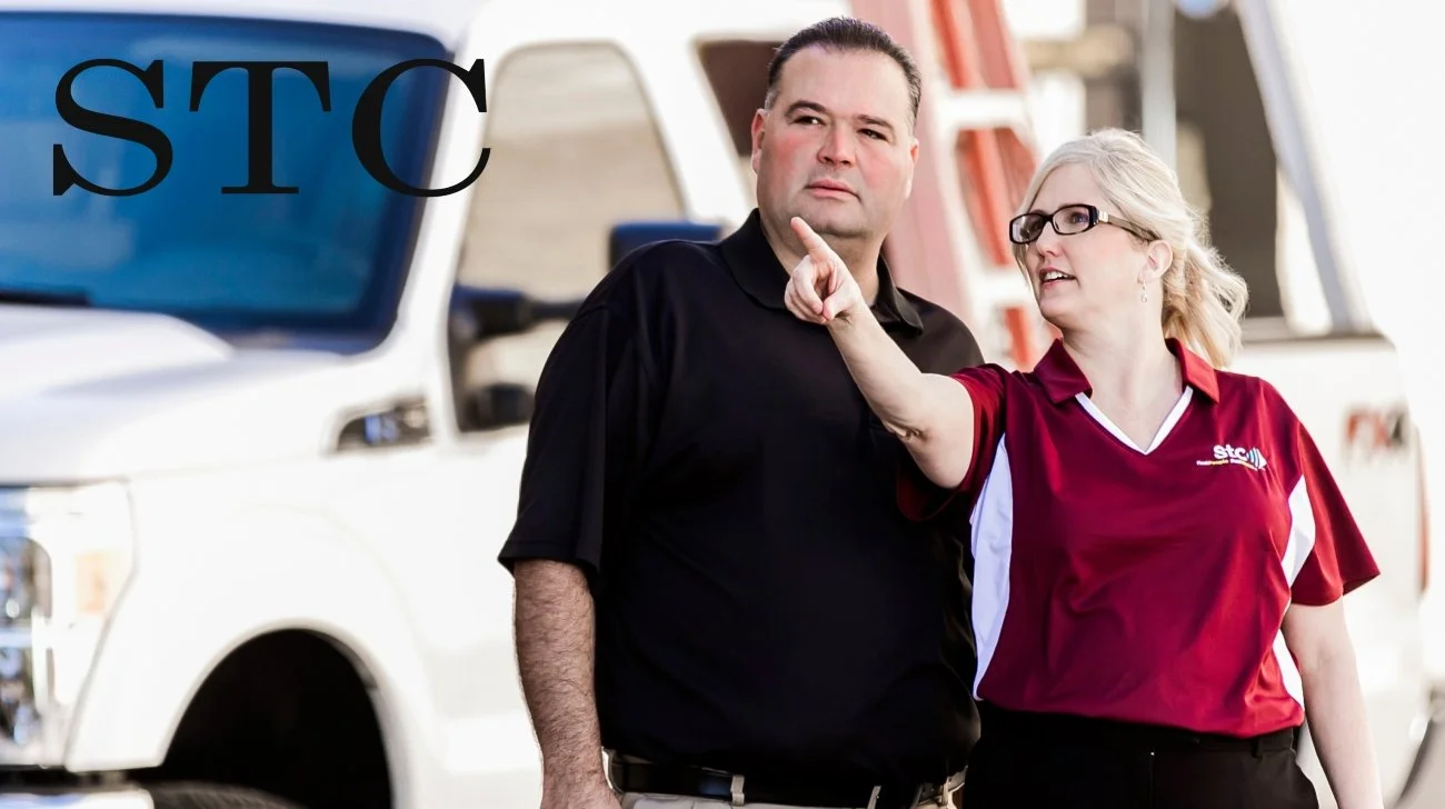 A man and a woman standing outdoors in front of a white utility truck. The woman, wearing a red shirt with a logo, is pointing and speaking to the man, who is dressed in a black shirt. The woman has blonde hair tied back and glasses, while the man ha