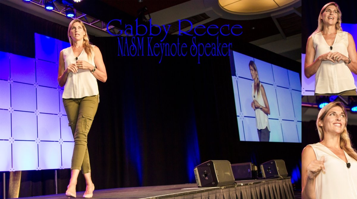 A woman standing on stage giving a keynote speech, with a large screen behind her displaying her image. The stage has black curtains and professional lighting.