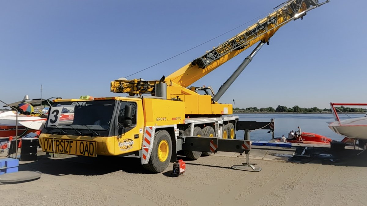 Yellow crane truck parked on sandy shore near water with boats, including a speedboat and a sailboat, under a clear blue sky.