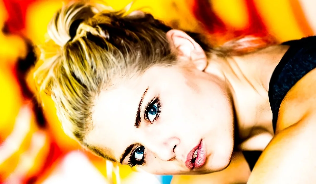Close-up of a woman with blonde hair lying on a colorful background, looking directly at the camera.