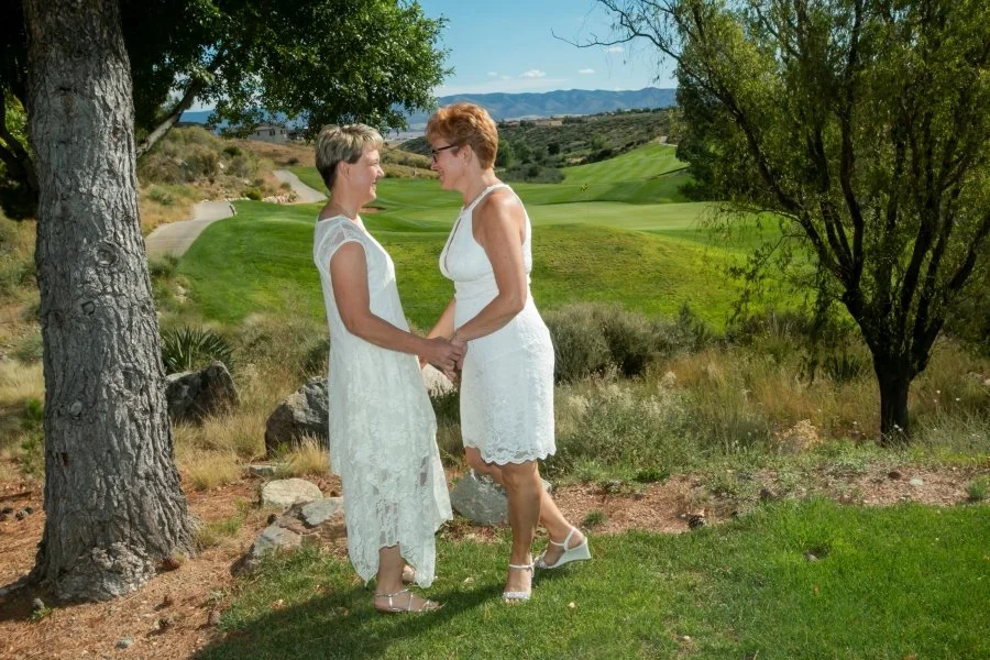 Two women in white dresses holding hands and smiling at each other outdoors on a grassy area with trees and hills in the background.