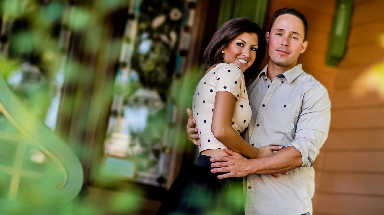 A smiling woman and a man hugging each other indoors with warm lighting and green decor in the background.