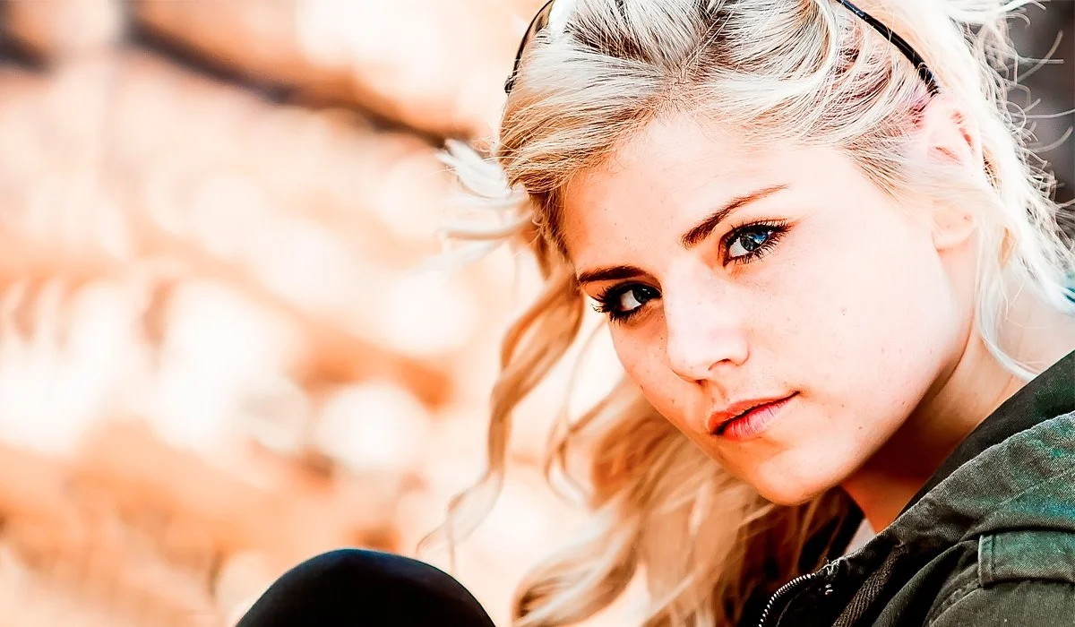 Close-up of a young woman with blonde hair and blue eyes, looking into the camera, outdoors with blurred background.
