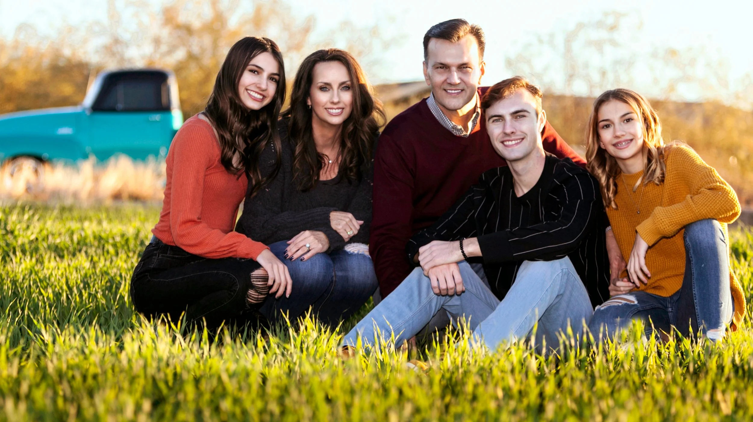 A family of six people, three women, two men, and a girl, sitting together on grass in an outdoor setting with trees and a blue truck in the background, during sunset or late afternoon.