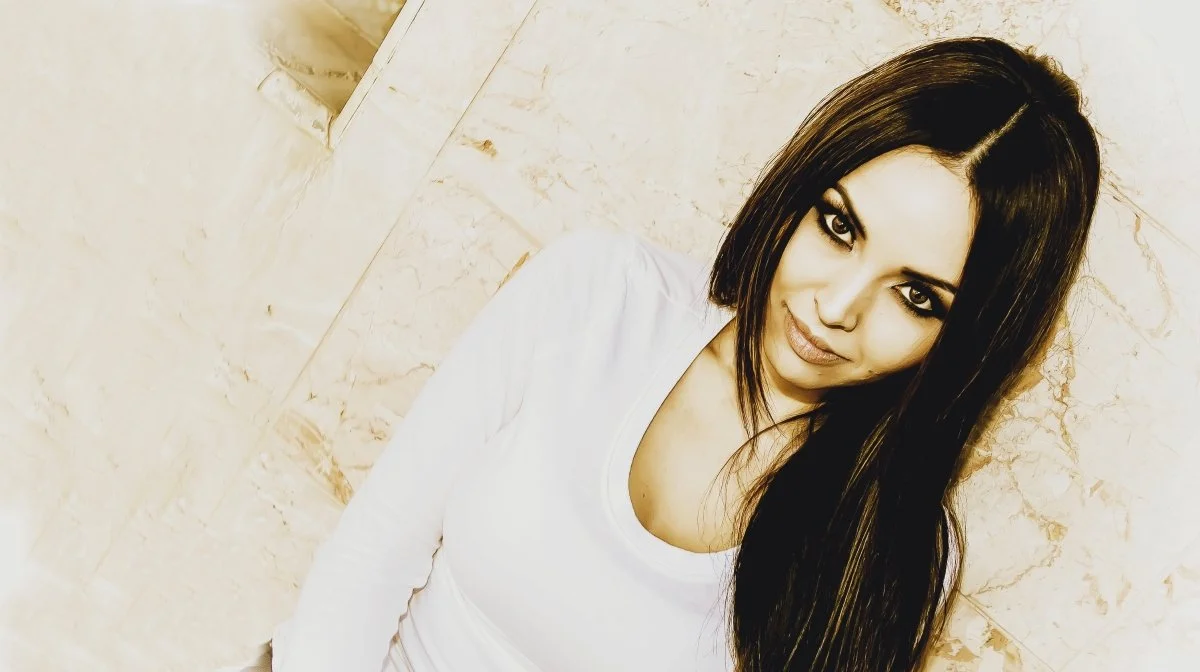 Portrait of a woman with long dark hair and makeup, wearing a white top, posing against a marble wall.