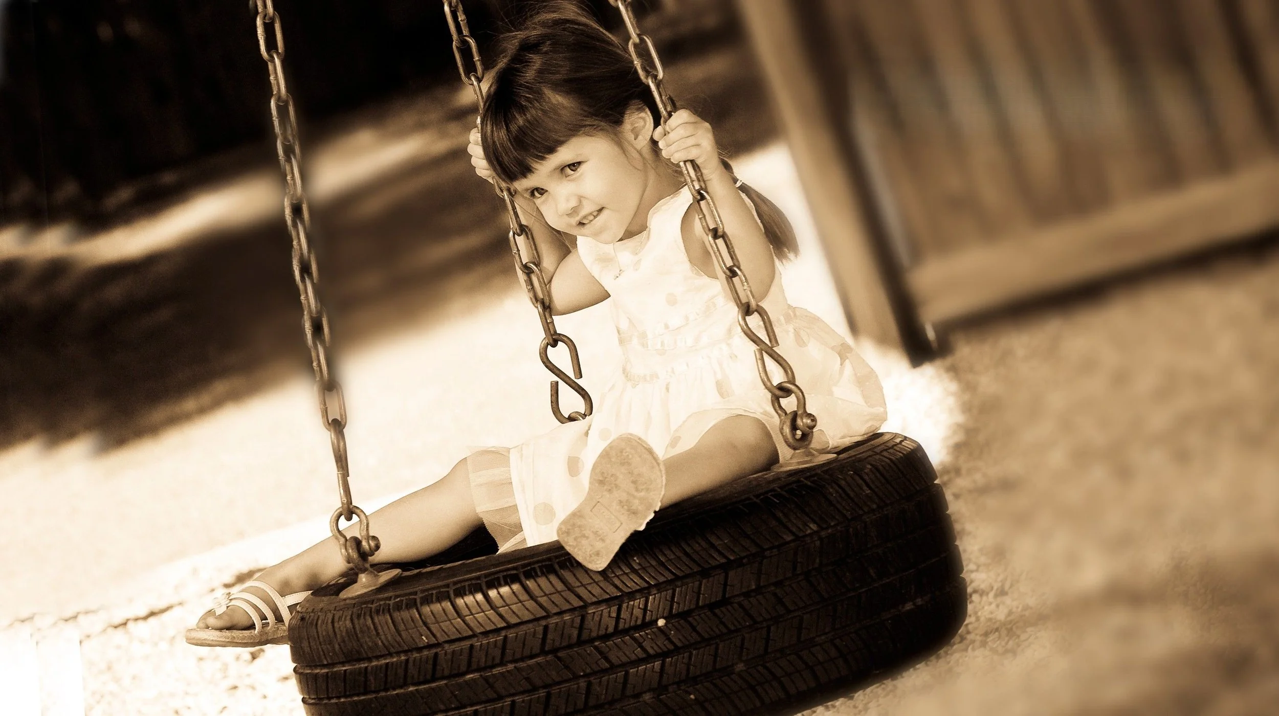 A young girl sitting on a tire swing at a playground, smiling and holding the chains, with a blurred background.