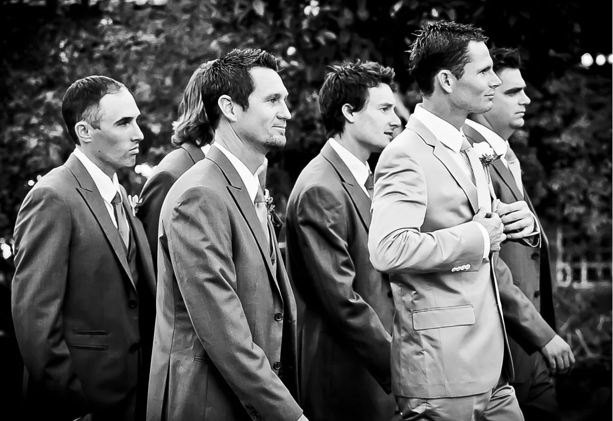 Seven men in suits standing in a row outdoors, appearing serious, with a background of trees.