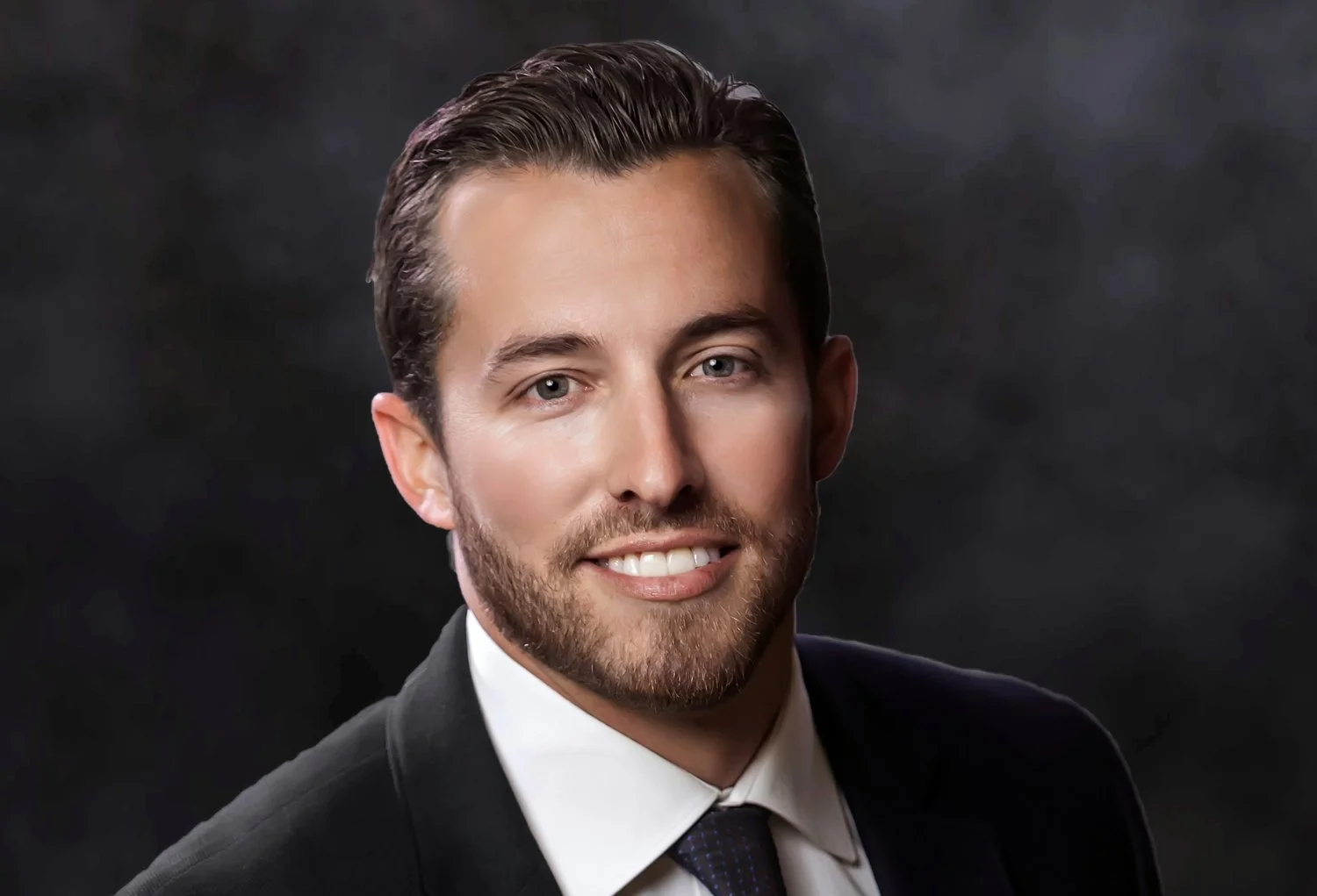 Headshot of a man with dark hair and a beard, wearing a black suit, white shirt, and dark tie, smiling against a dark background.