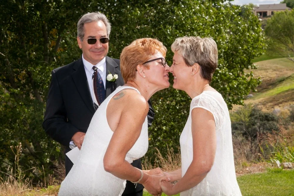 Two women in wedding dresses holding hands face-to-face in an outdoor setting, with a man in a suit and sunglasses smiling in the background.