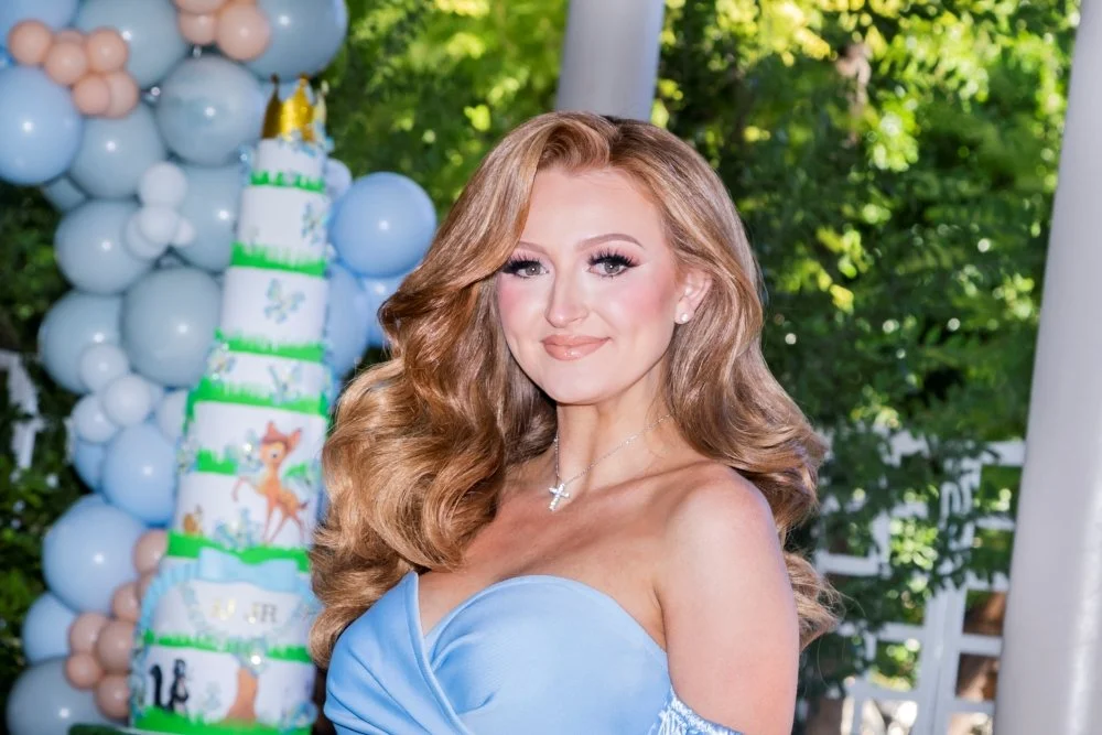 A woman with long, wavy red hair and fair skin, wearing a light blue strapless dress, standing outdoors near a decorated birthday cake with a forest and balloons in the background.