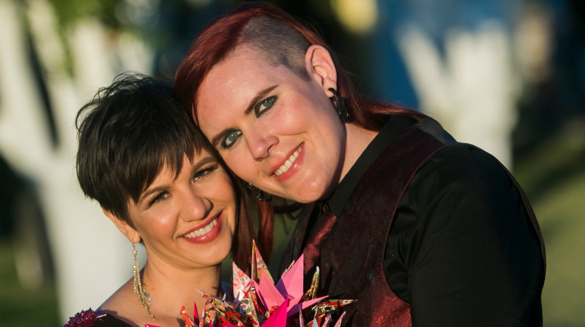Two women smiling, one with short dark hair and the other with shoulder-length red hair, embracing and holding a pink floral bouquet outdoors.