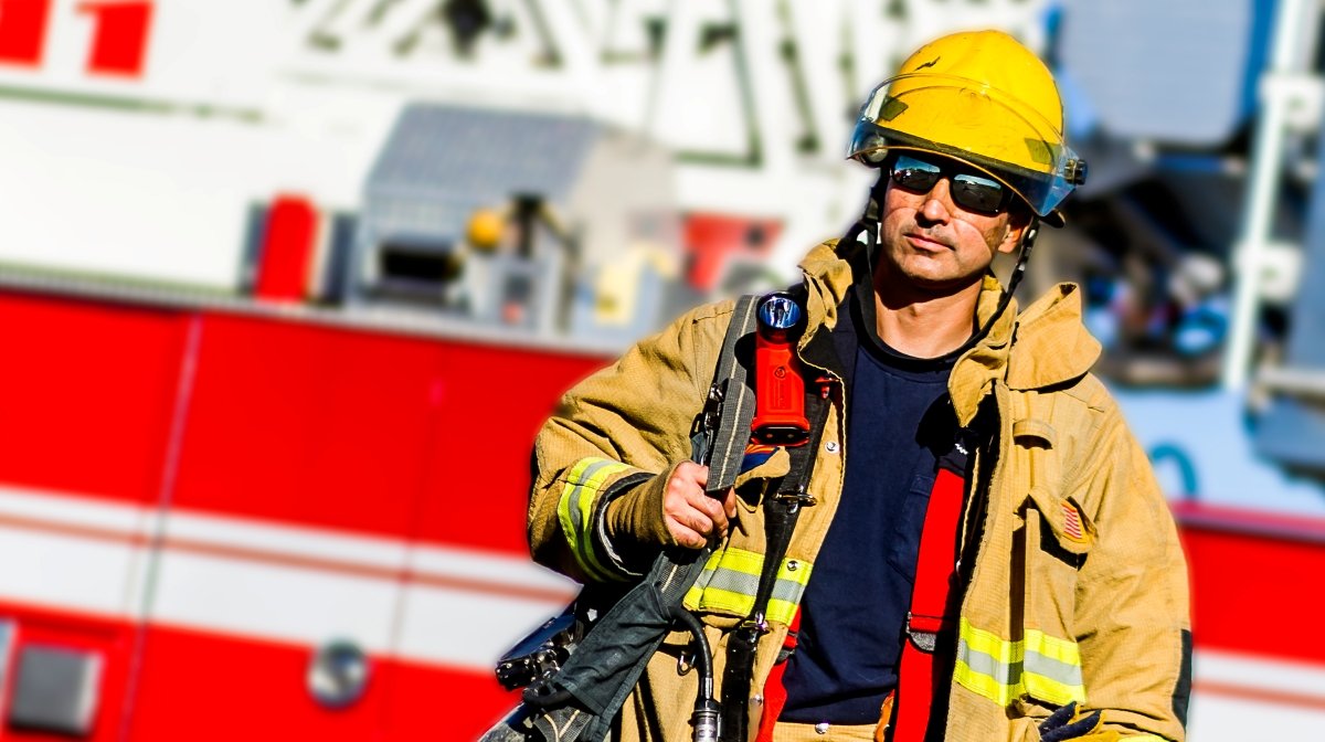 A firefighter wearing a yellow helmet, sunglasses, and tan turnout gear standing in front of a red fire truck.