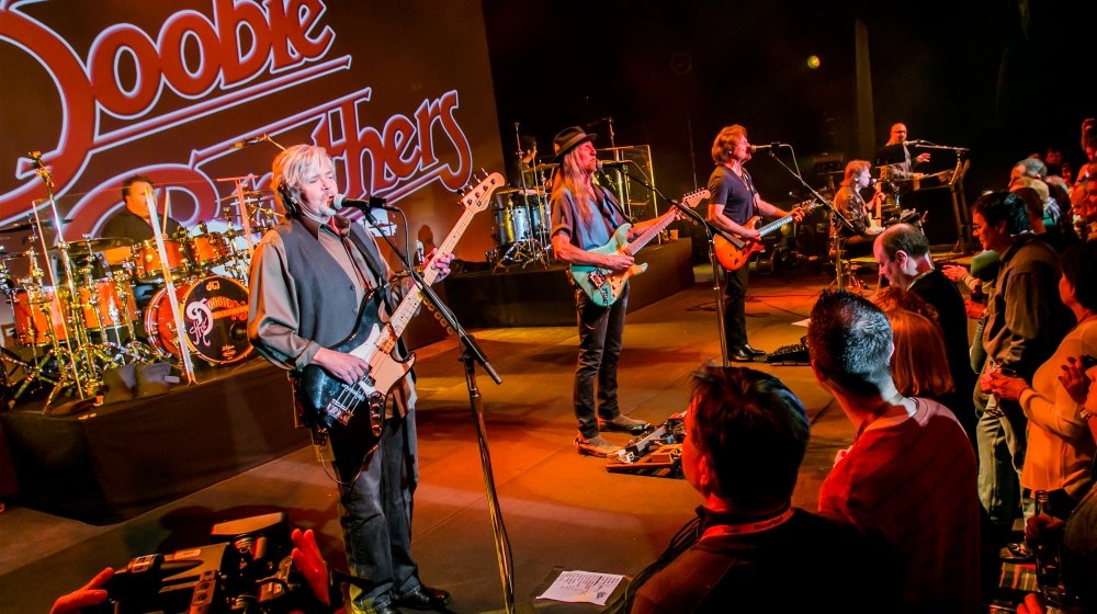 A band performing live on stage with a sign that reads "Doodoo and the Butter" in the background, including a drummer, several guitarists, and a keyboard player, with an audience watching.