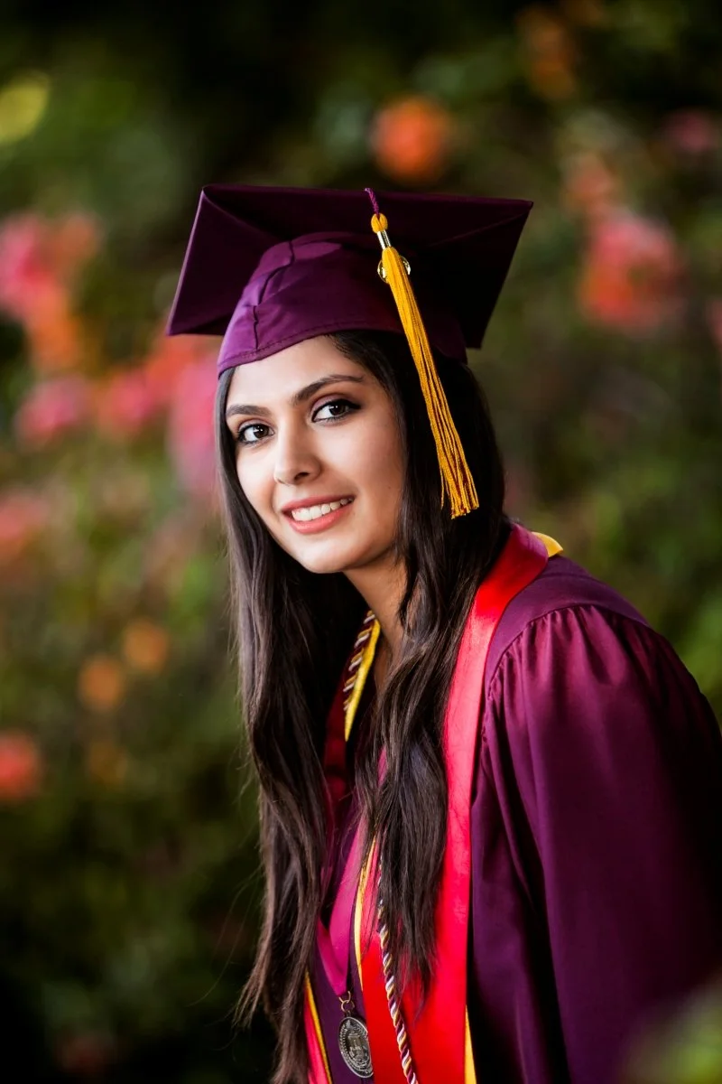 A young woman in a maroon graduation gown and cap with a gold tassel, smiling outdoors with a colorful, blurred leafy background.