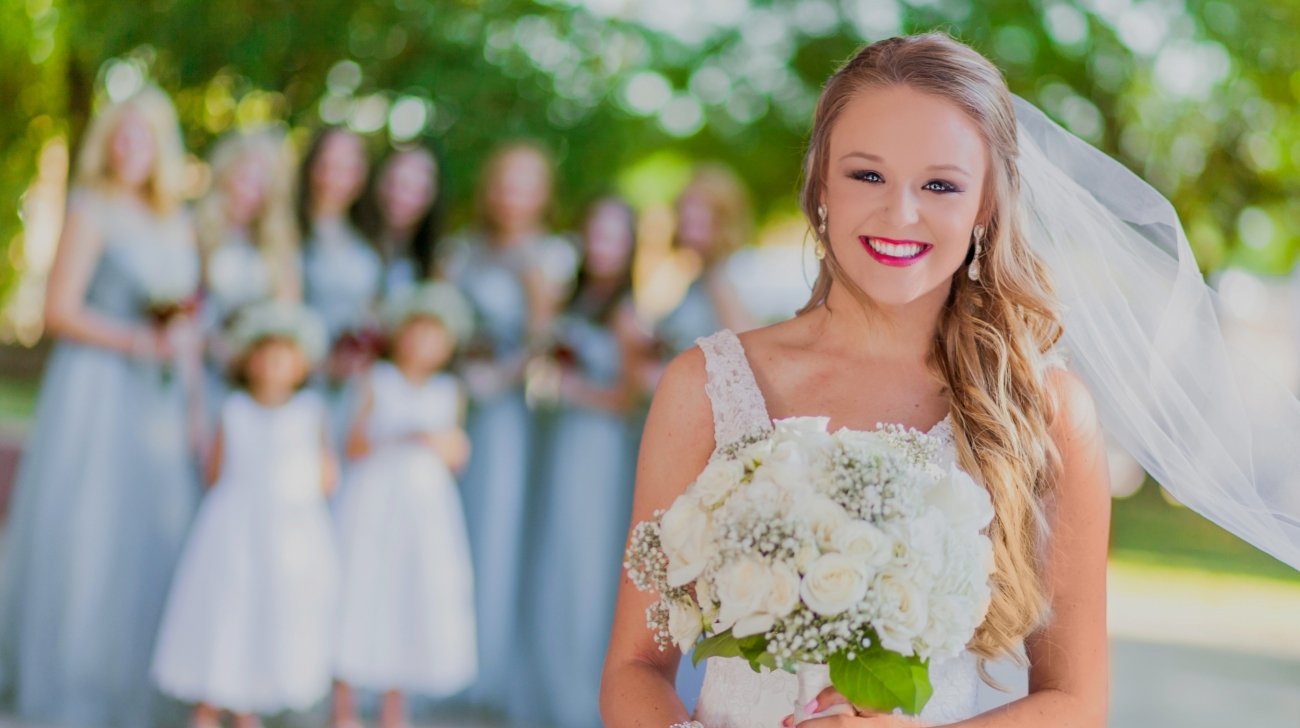 Bride smiling and holding a bouquet of white flowers, with bridesmaids and flower girls in matching dresses blurred in the background outdoors.