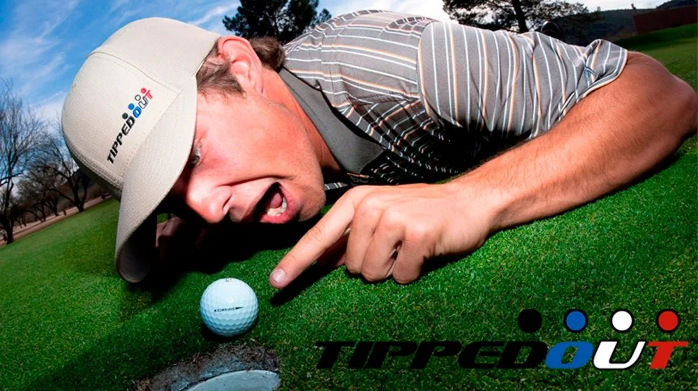 A man lying on a golf course grass, reaching towards a golf ball with a golf hole nearby, wearing a beige cap and a striped shirt, with the logo 'TIPPED OUT' at the bottom.