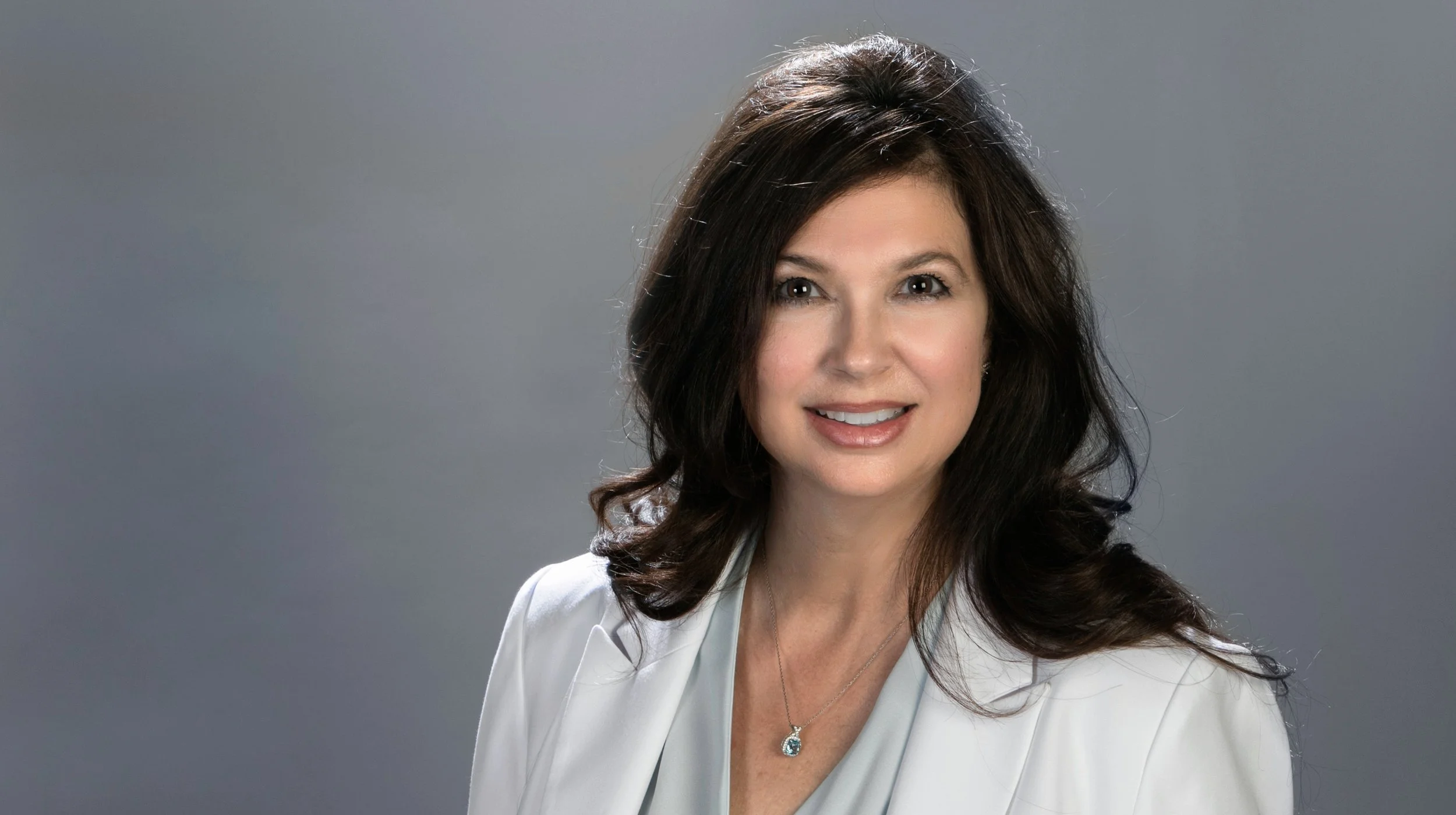 Professional woman with dark brown hair, smiling, wearing a white blazer and a silver necklace with a pendant, against a plain gray background.