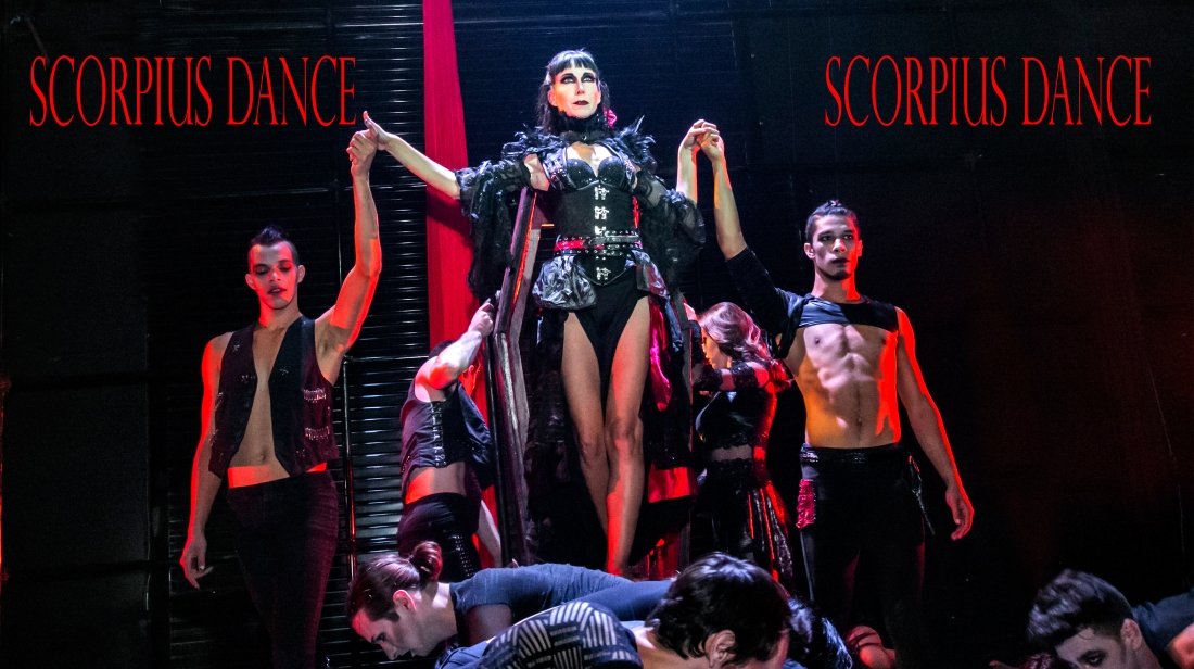 Performance of Scorpius Dance featuring a woman in a dramatic costume on stage with backup dancers, with black and red backdrop and the words 'Scorpius Dance' displayed.