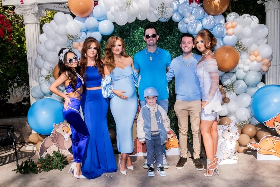 Group of six people, including a pregnant woman, posing in front of a balloon arch with cartoon animal cutouts, celebrating a baby shower or gender reveal outdoors.