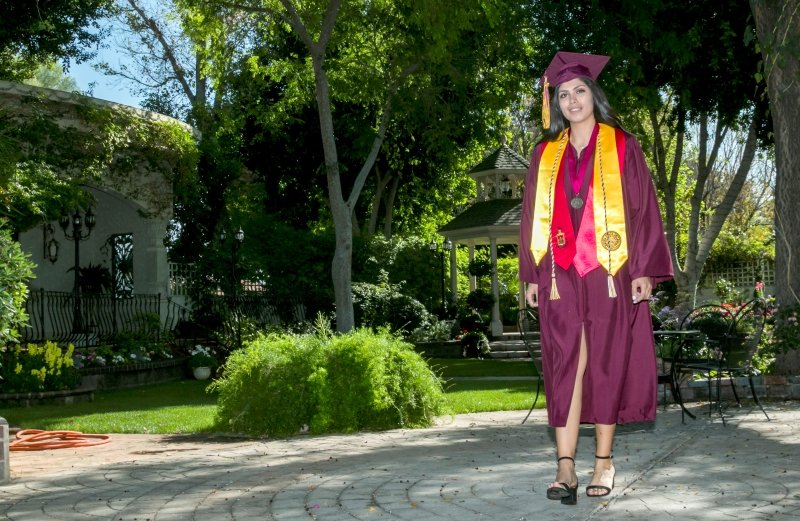 A young woman in a maroon graduation gown and cap stands outdoors on a paved area, smiling. She has a yellow honor cord and a medal around her neck. The background features green trees, a gazebo, and garden furniture.