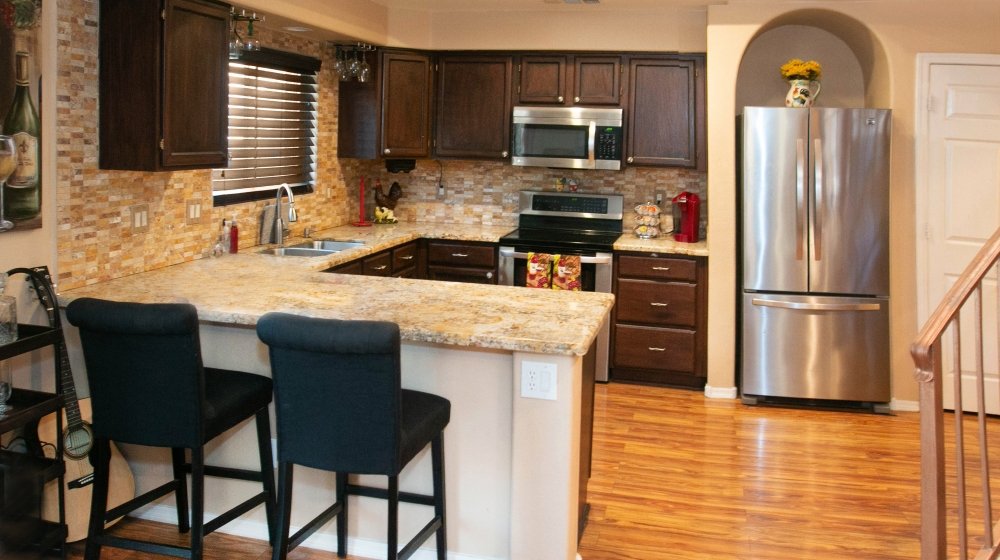 Kitchen with dark wood cabinets, granite countertops, a stainless steel refrigerator, stovetop, microwave, and a window with blinds. There are two black barstools at the counter and hardwood floors.
