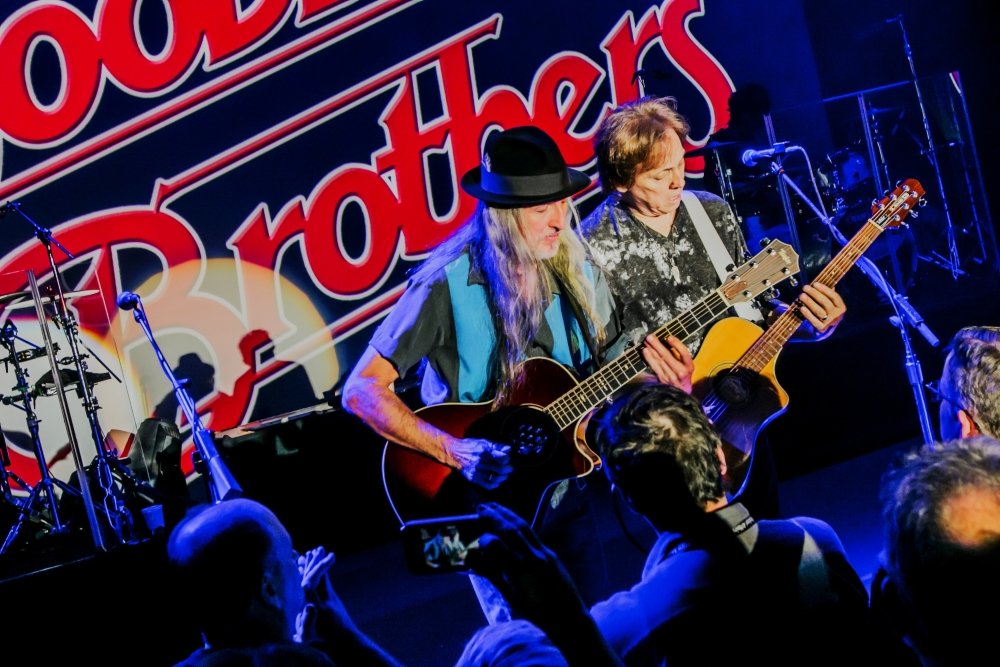 Two musicians playing guitars on stage during a live concert, with an audience watching in the foreground, and a large red and white sign in the background.