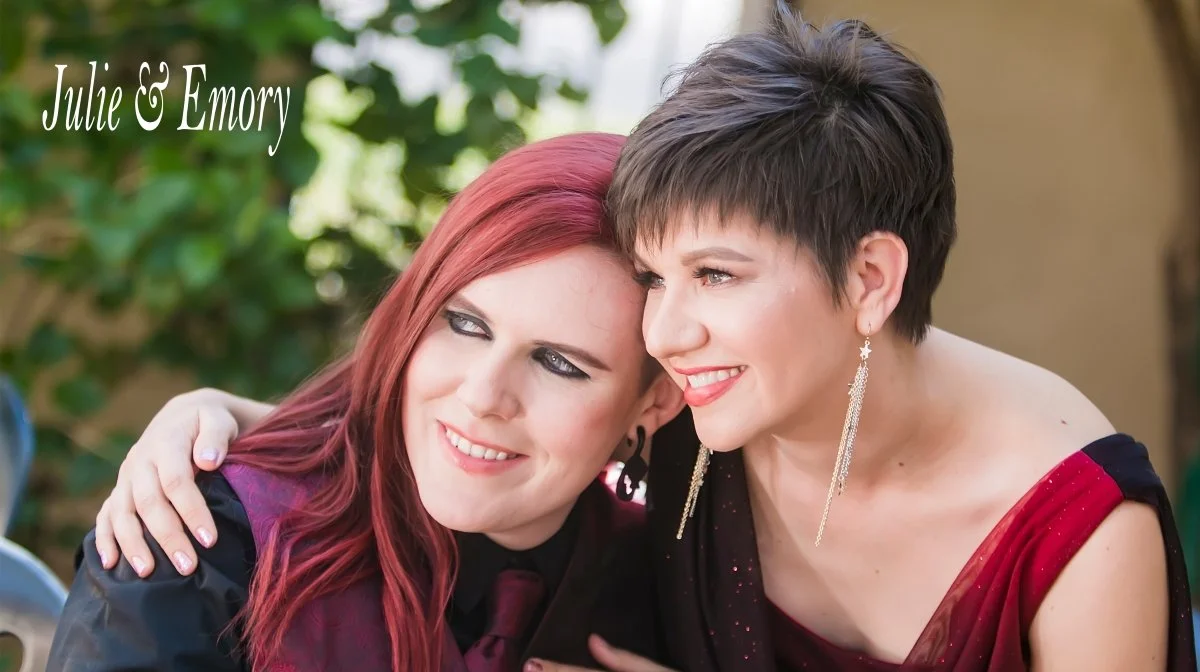 Two women with short dark hair and long red hair smiling and hugging outdoors, with greenery in the background, wearing elegant dresses and jewelry, celebrating together.