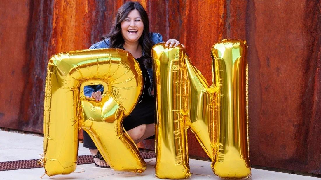 Woman smiling while holding large gold balloon letters spelling "RMI" in front of a rust-colored wall.