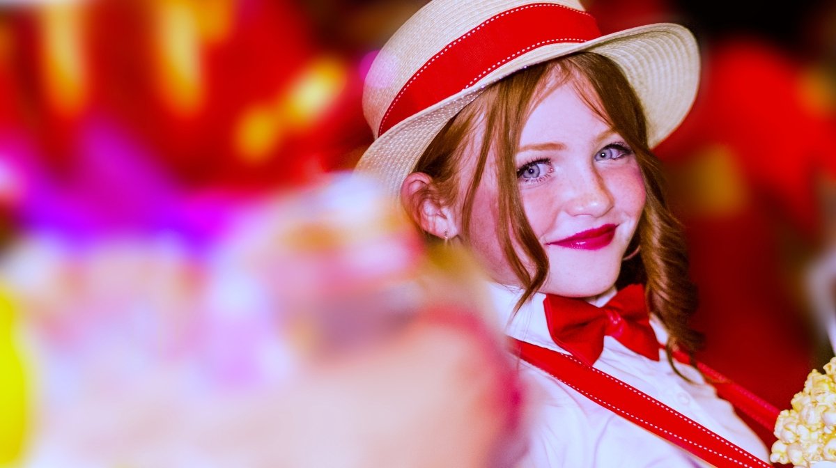 Young girl with red hair, wearing a straw hat with red ribbon, white shirt, and red bow tie, smiling in a colorful setting.