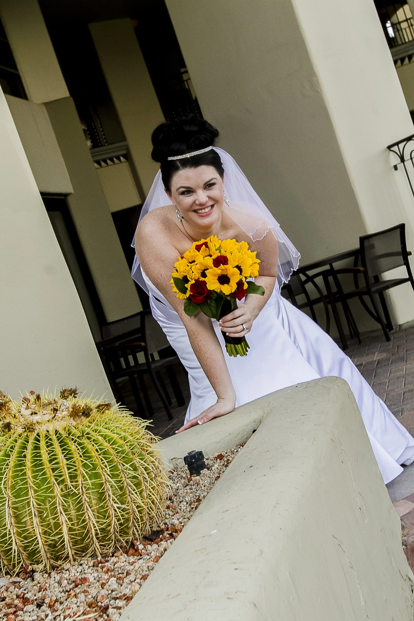 A bride in a white wedding dress leaning on a low concrete wall, holding a bouquet of yellow and red flowers, smiling at the camera.