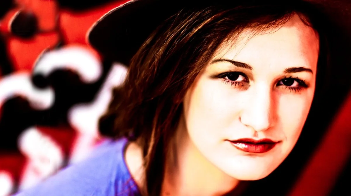 A young woman with dark brown hair, wearing a black hat and purple top, looking directly at the camera with a slight smile, in front of a blurred colorful background.
